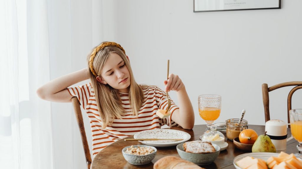 Teen girl in striped shirt at breakfast table with fruits, bread, and juice. Casual morning setting.