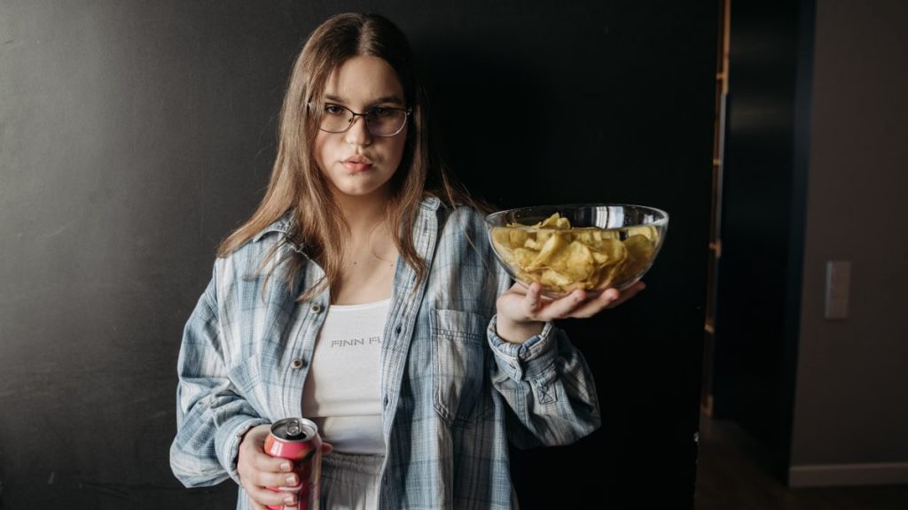 Casual woman holds chips and soda, posing indoors against a dark background.